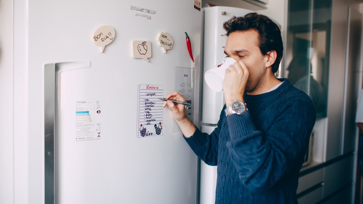 Young male employee in casual outfit drinking coffee and taking notes on paper attached on fridge in morning
